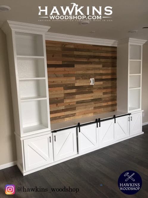 Front view of a handcrafted built-in media wall and bookcase unit with reclaimed wood plank center panel, black barn door hardware, and two white bookcase towers with crown molding, made to order in the USA by Hawkins Woodshop